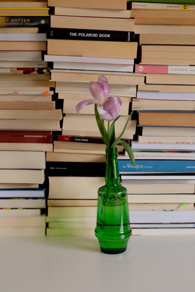 A stack of books with a pink flower in a green glass vase